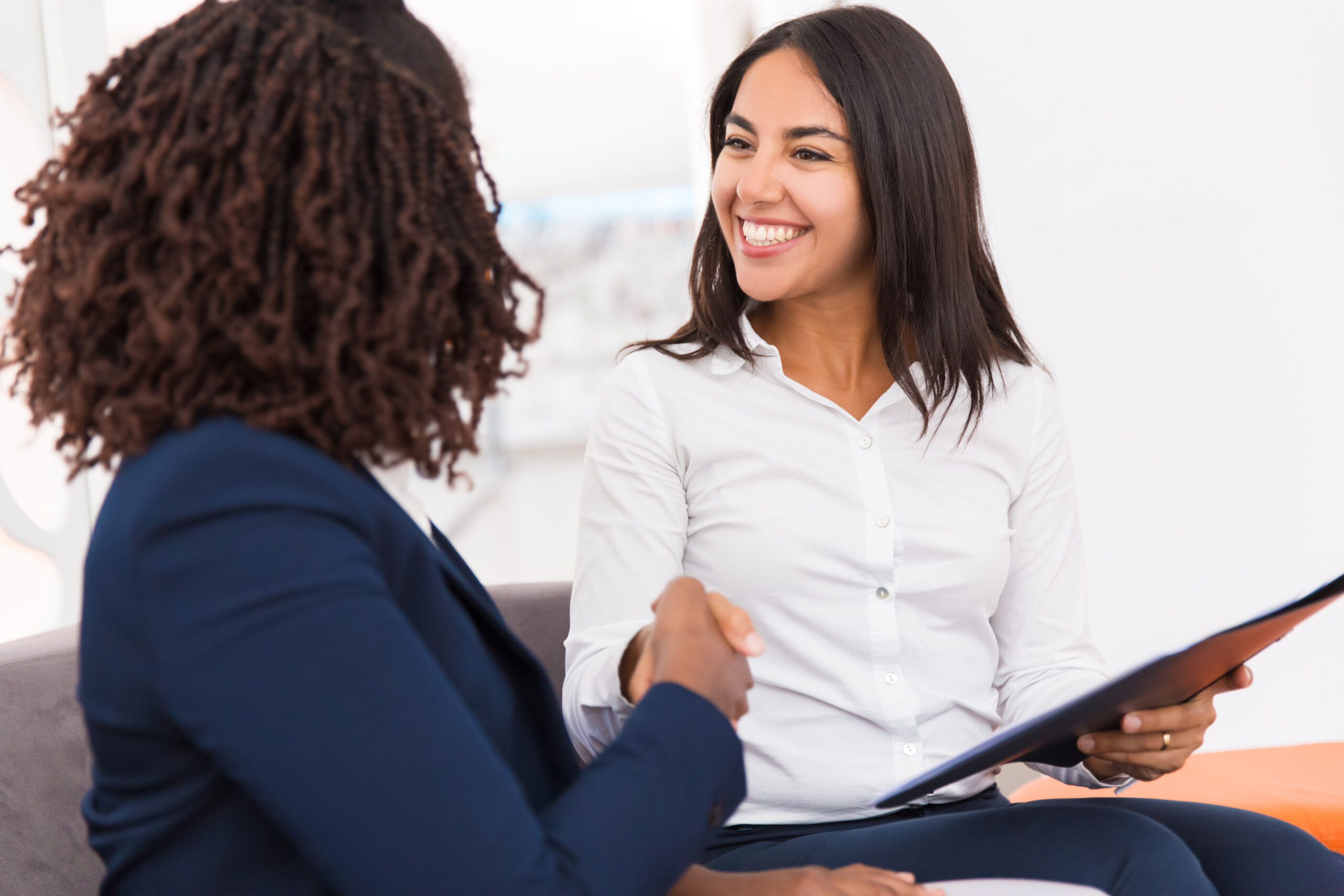 Happy diverse business partners closing deal. Businesswomen sitting on couch, holding folder with documents, shaking hands and smiling. Agreement concept